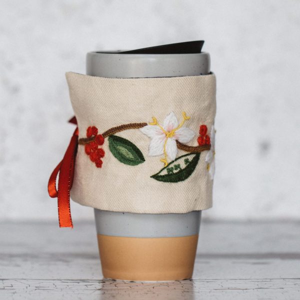 Travel coffee mug in gray ceramic and wood, covered. with a linen in a crewel work design with ribbon tie.